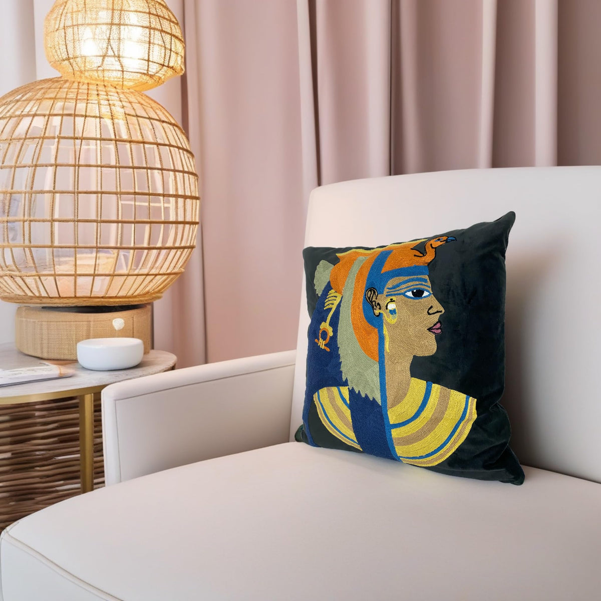 Embroidered velvet cushion cover with a Cleopatra portrait on a cream armchair by a rattan lamp