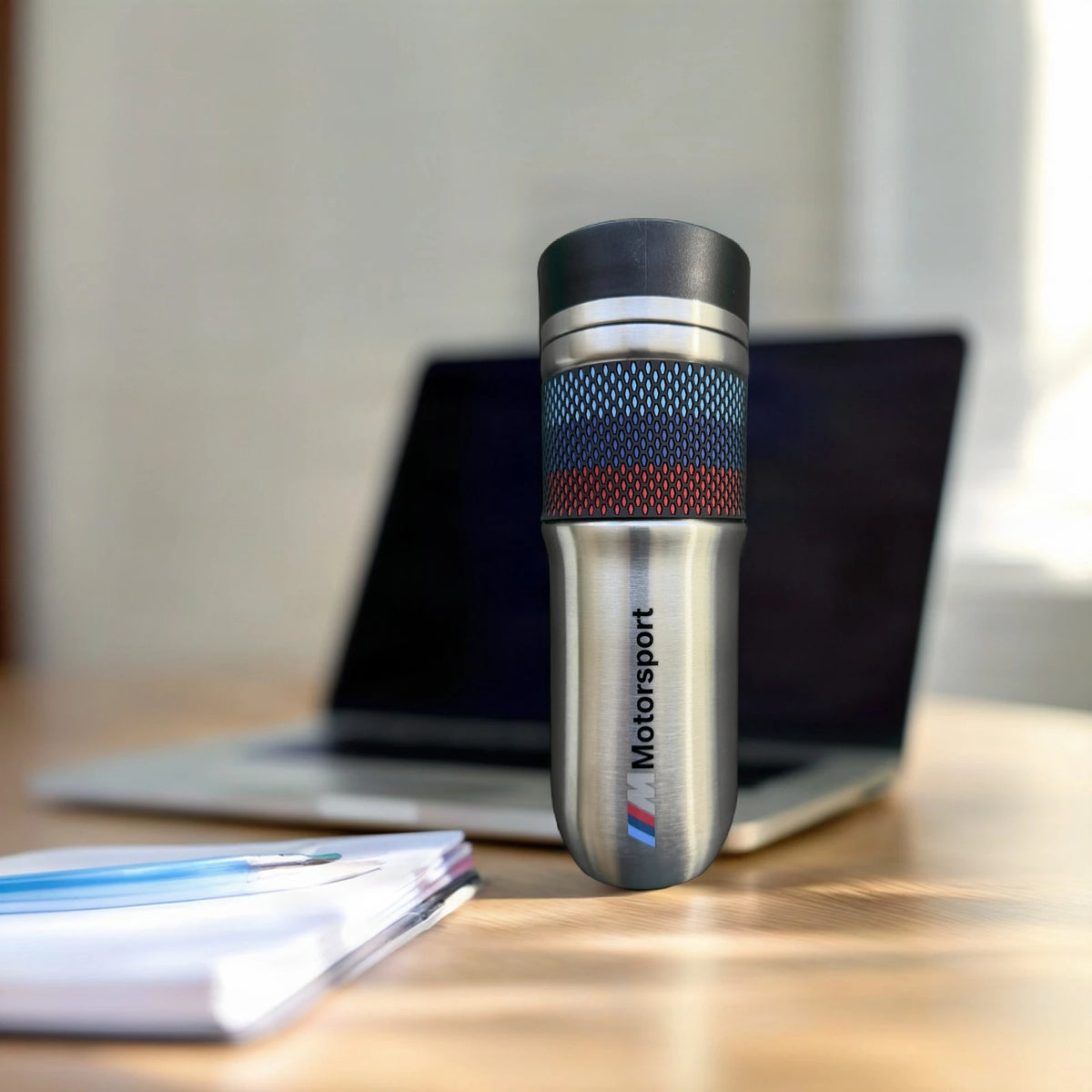 BMW thermos travel mug on an office desk beside a laptop, M Motorsport wordmark facing forward