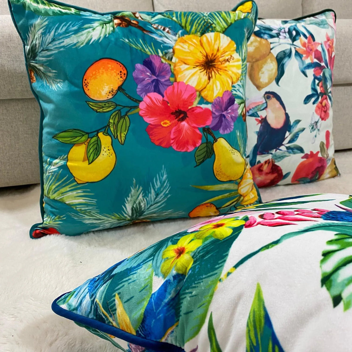Teal tropical, toucan and floral velvet cushion covers layered on a cream sofa and sheepskin rug