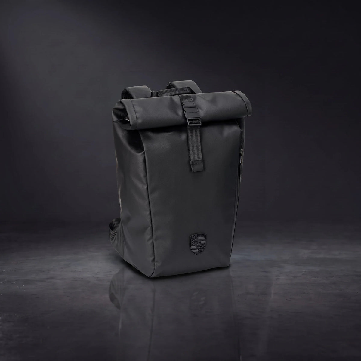 Black Porsche 911 roll-top backpack standing on a glossy dark studio floor, front view with the 3D crest