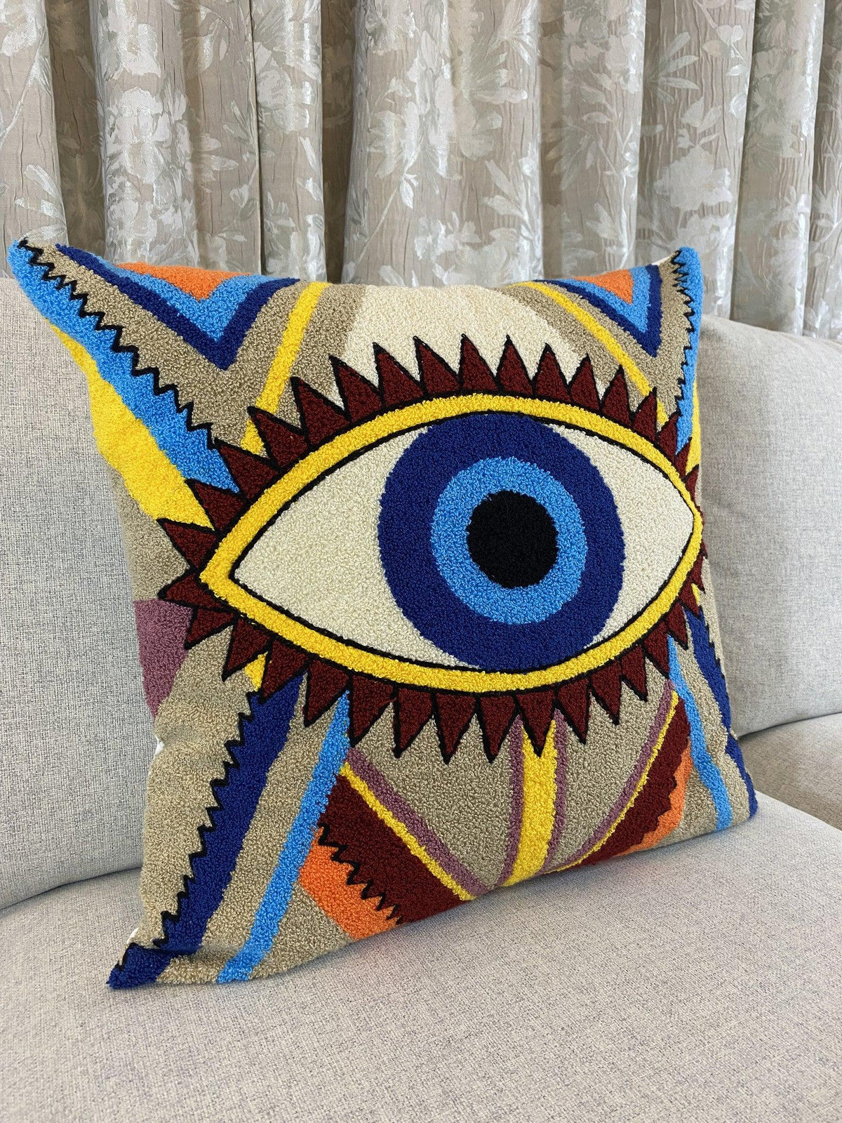 Tufted 100% cotton evil eye cushion cover standing on a grey sofa in front of curtains