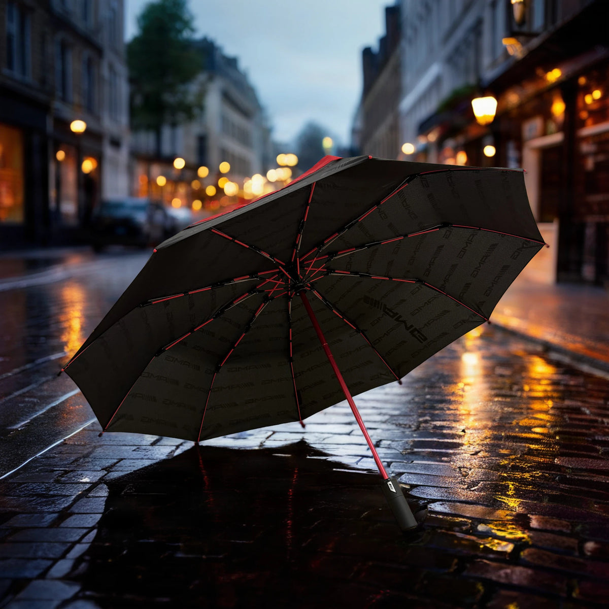 Mercedes-AMG black and red folding travel umbrella open on wet cobblestones on a rainy London street at dusk