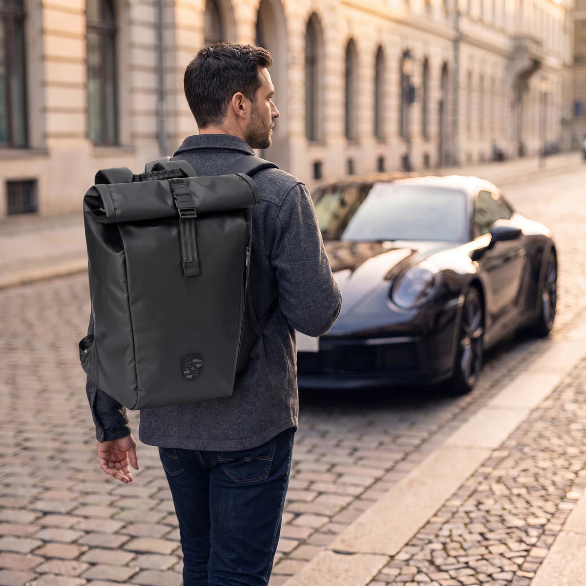 Man wearing the black Porsche 911 roll-top backpack on a cobbled city street beside a parked black sports car