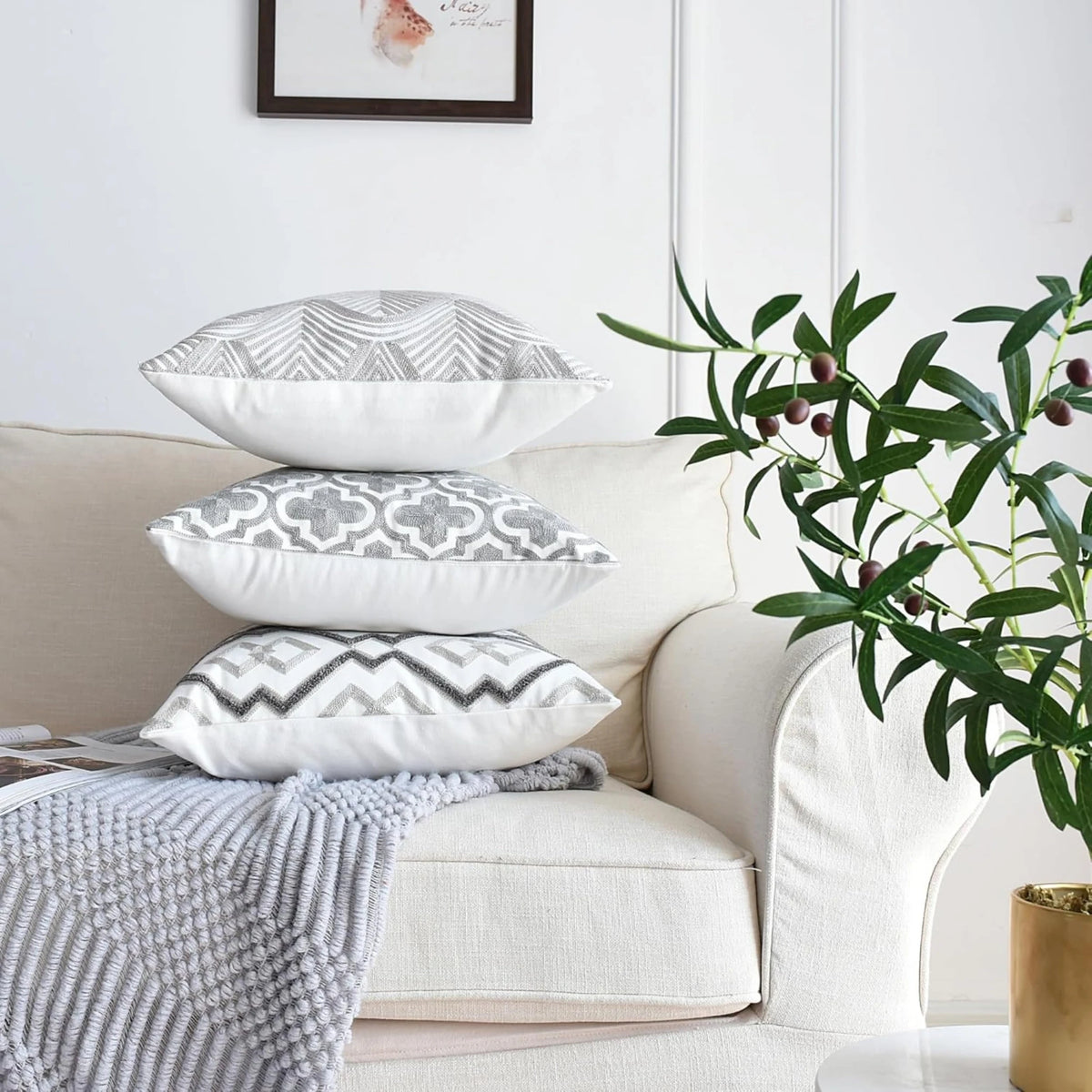 Three grey and white embroidered cushion covers from the set of 4 stacked on a sofa arm beside an olive branch